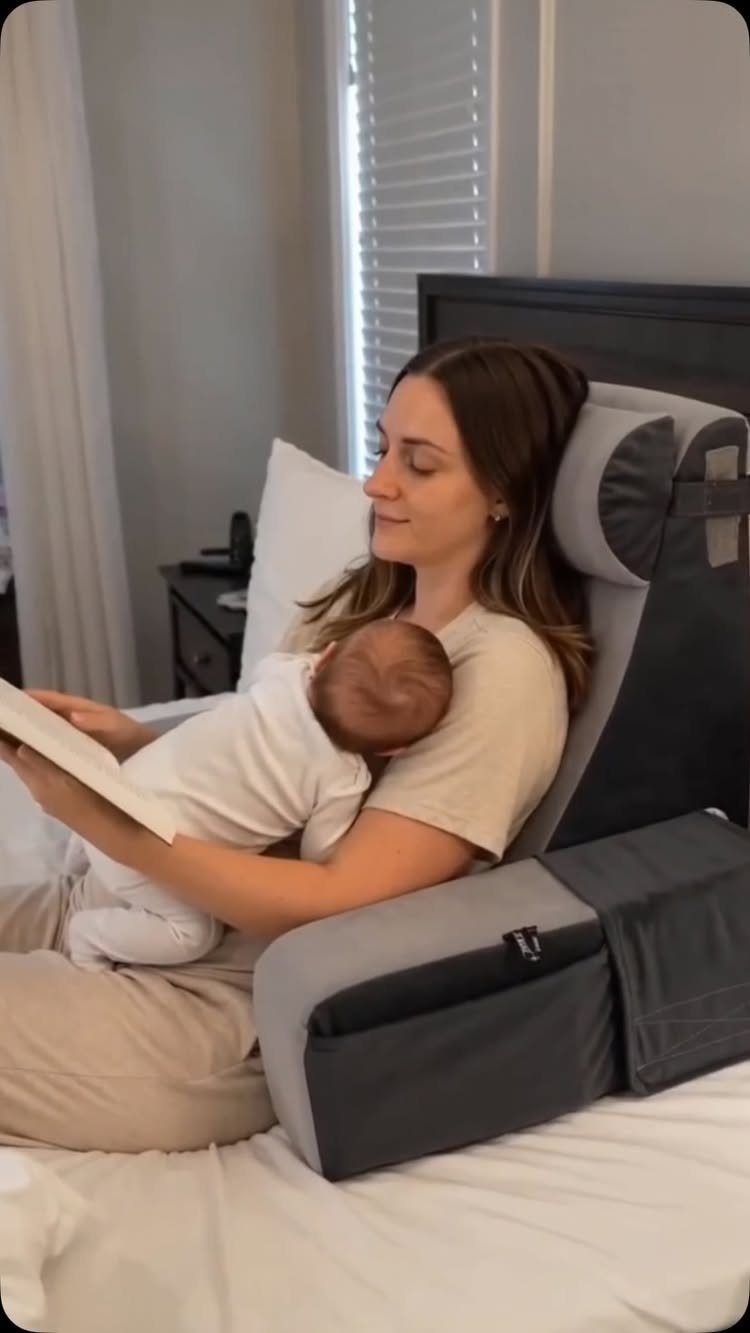 Woman sitting in bed with a baby, using a grey nursing pillow for support.