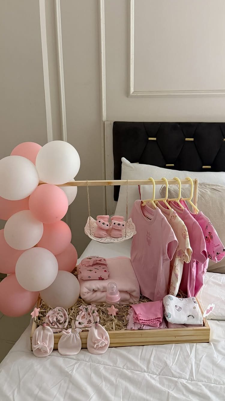 Wooden gift tray with pink baby clothes, blankets, and pink and white balloons.