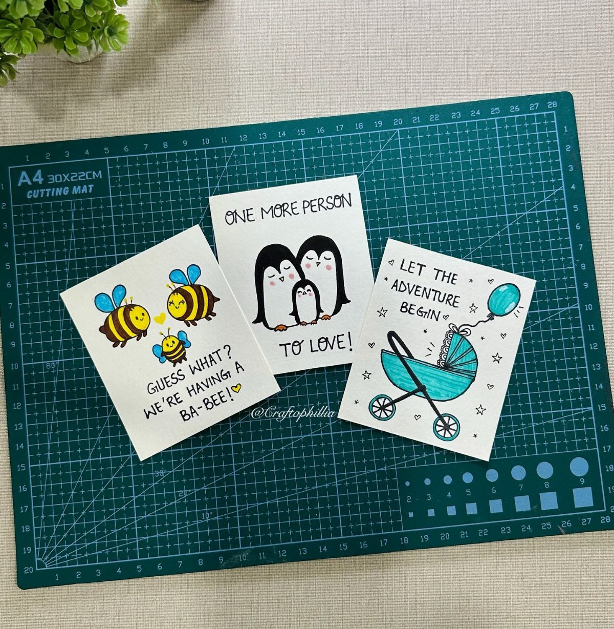 Three hand-drawn pregnancy announcement cards resting on a green cutting mat.