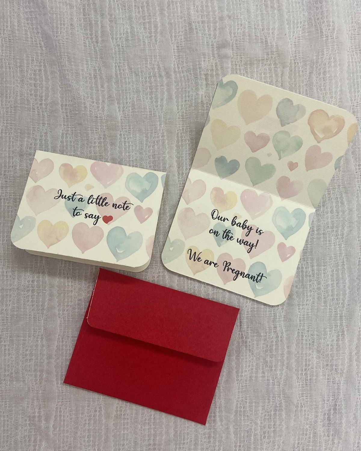 Two pregnancy announcement cards with pastel hearts and a red envelope on fabric.