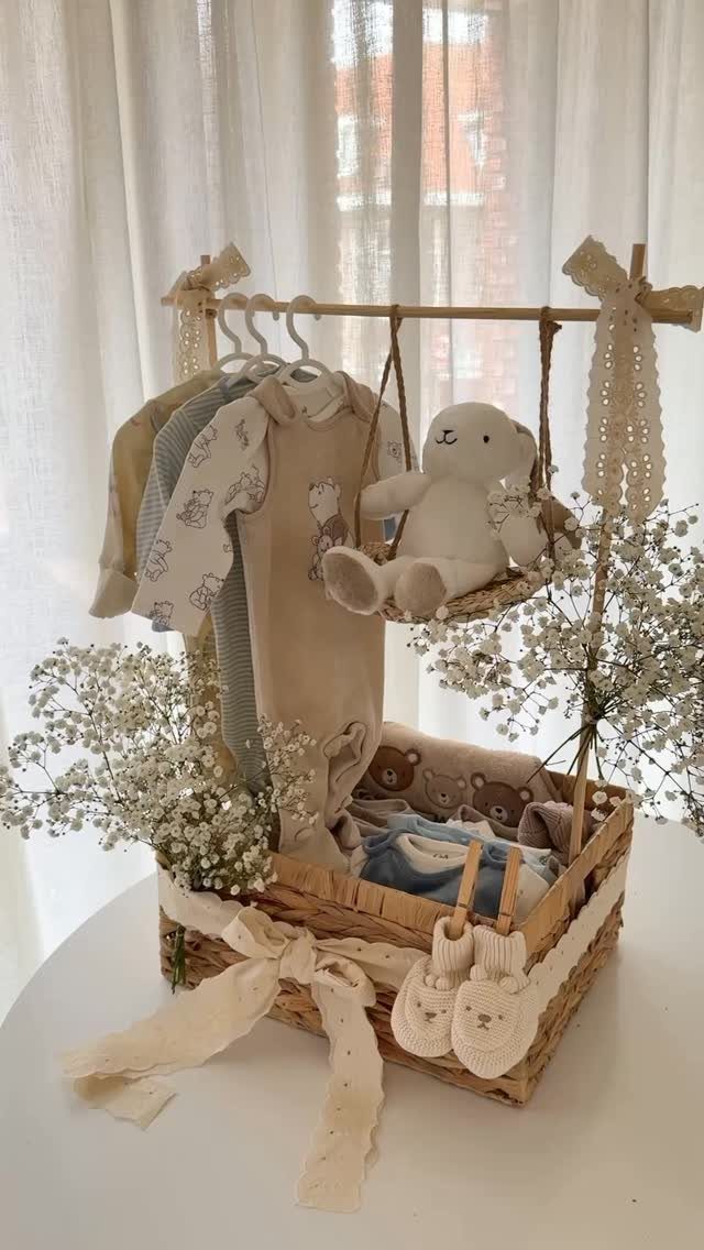 Woven wicker gift basket containing baby clothes, a teddy bear, and white flowers.