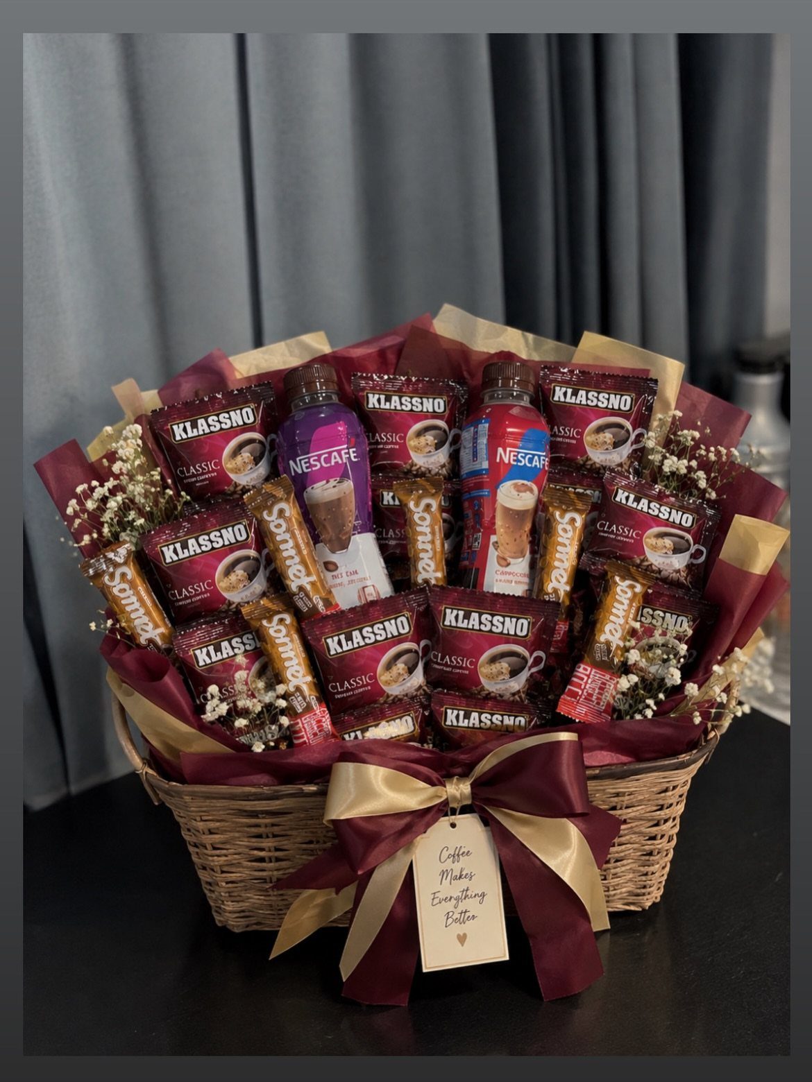 Wicker gift basket filled with coffee products, burgundy ribbon, and white flowers.