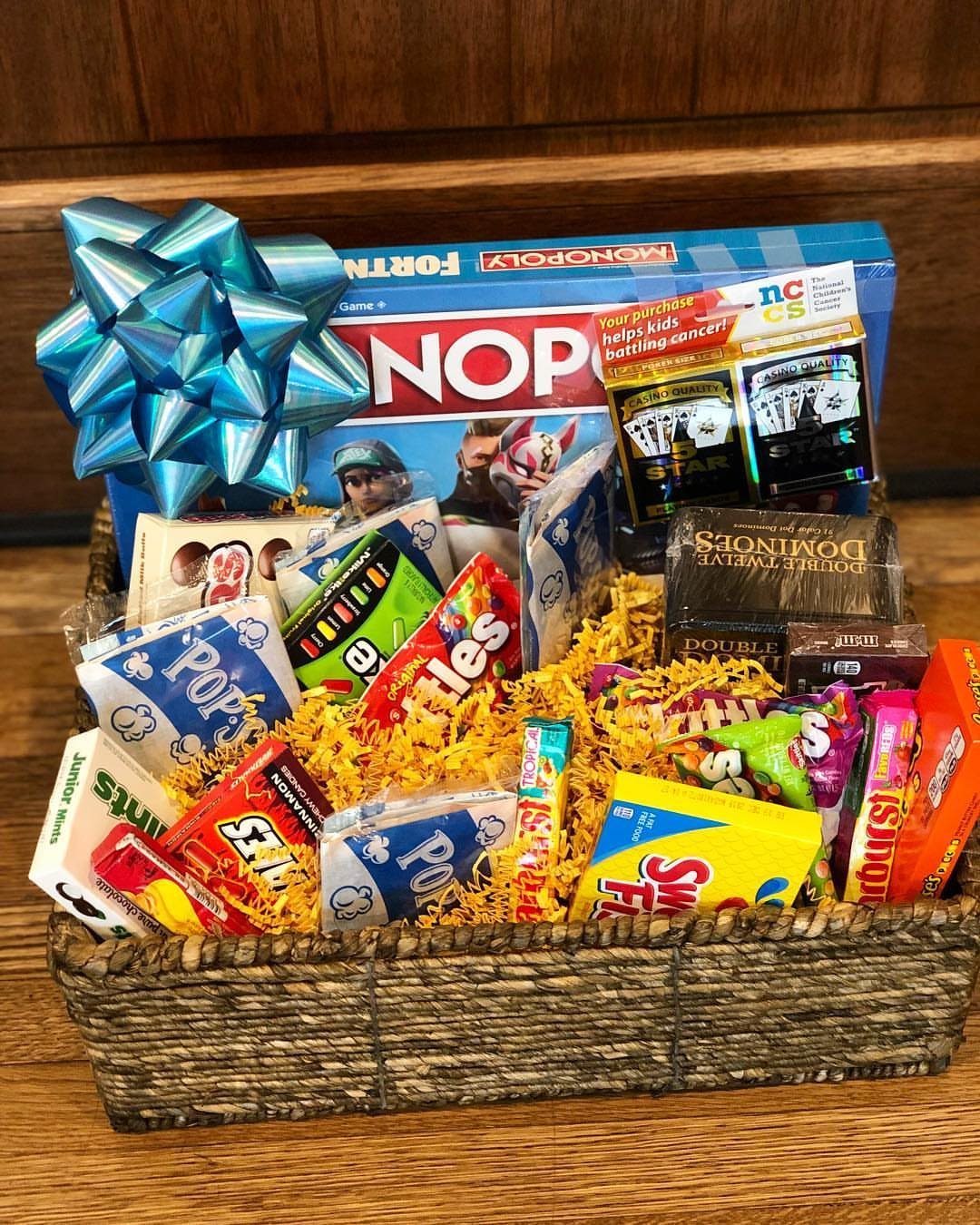 Woven gift basket with Monopoly board game, dominoes, and assorted colorful snacks.