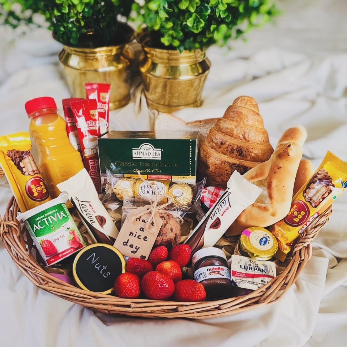 Breakfast basket with croissant, baguette, orange juice, strawberries, and various spreads on white cloth.