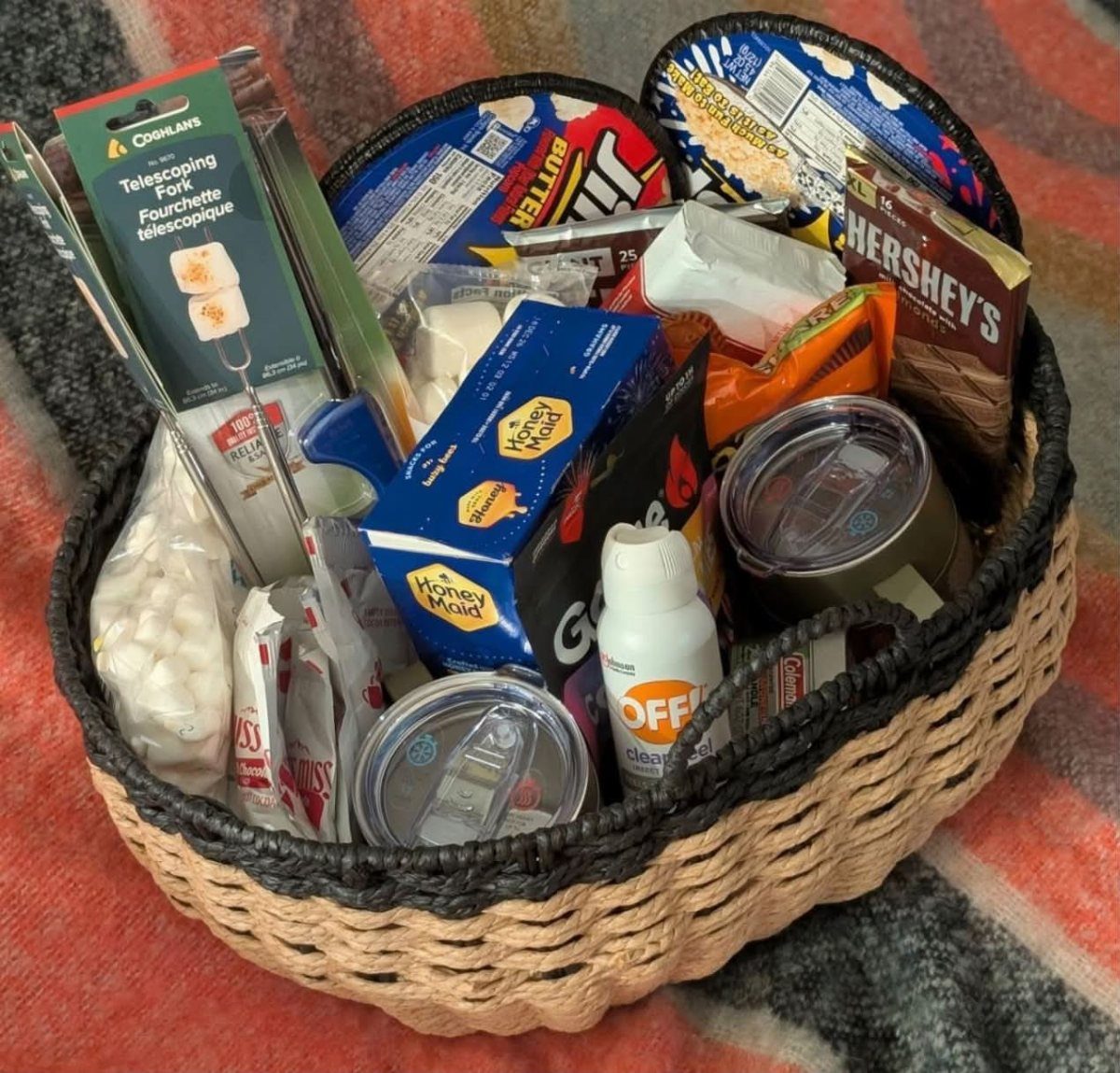 Woven basket filled with s'mores ingredients, bug spray, and camping gear on a blanket.