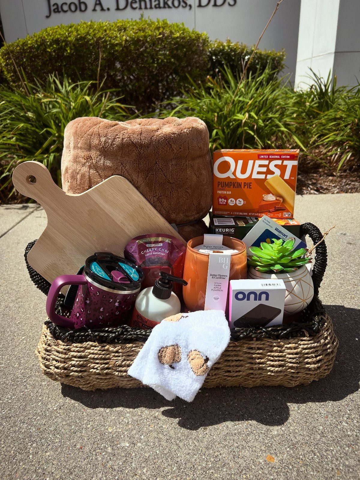 Woven gift basket containing a cutting board, plush towel, mug, and protein bars.
