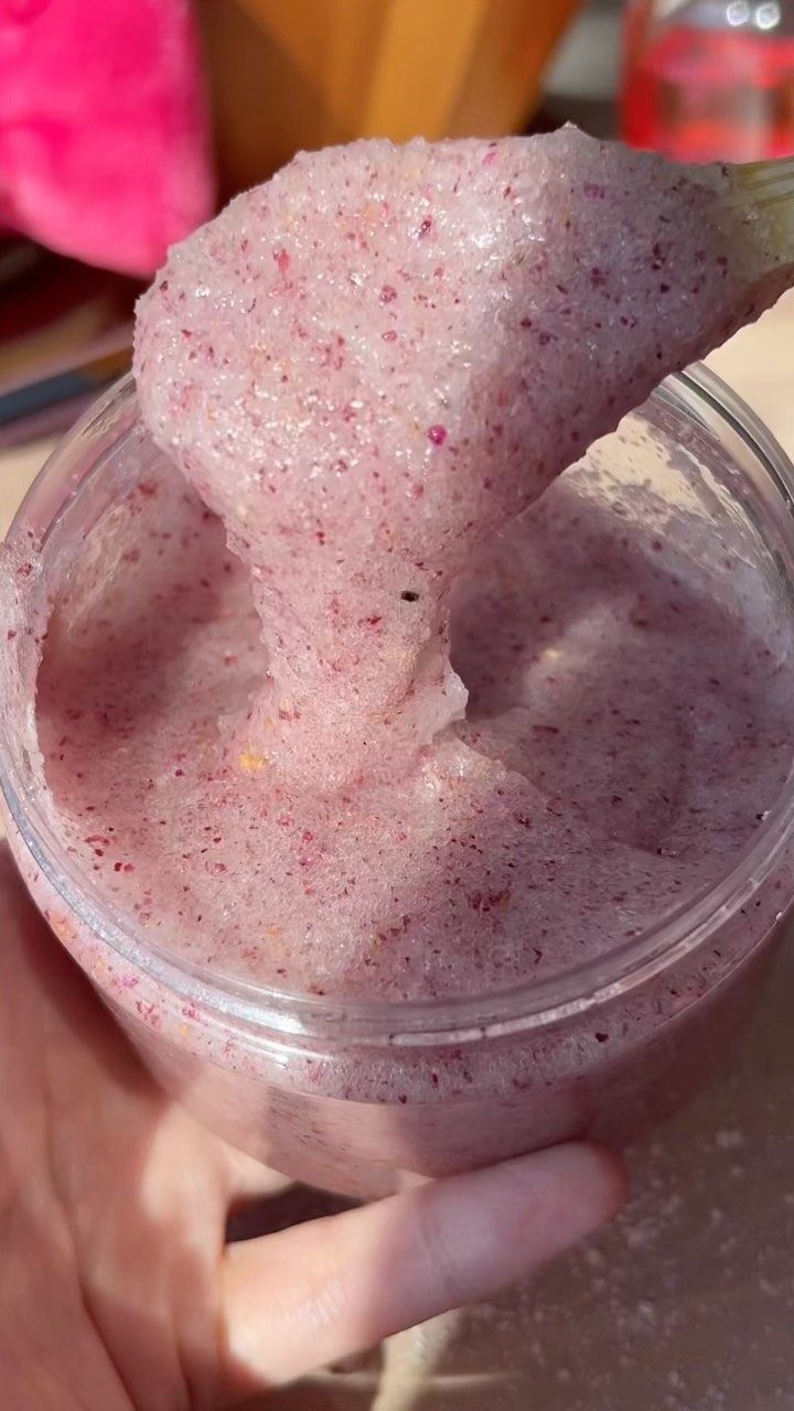Pinkish-purple sugar scrub being scooped out of a clear plastic jar with a spoon