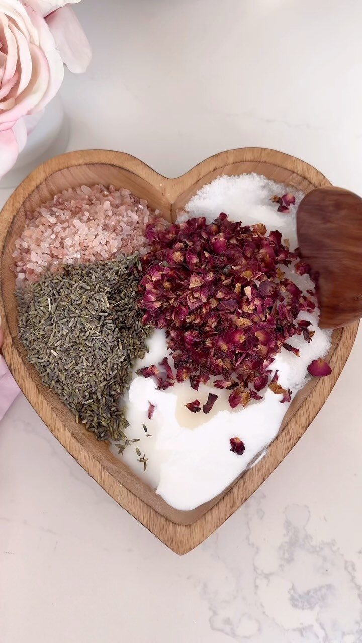 Heart-shaped wooden bowl filled with pink salt, white salts, herbs, and rose petals.