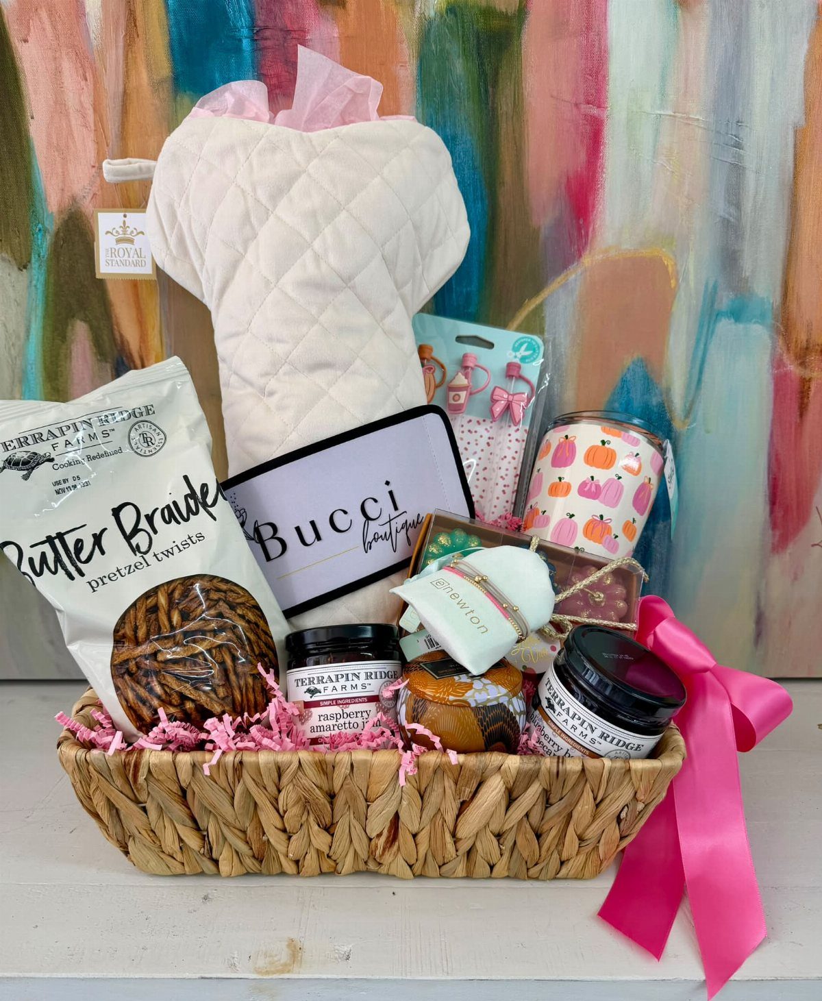 Woven gift basket filled with a white quilt, snacks, and jams with pink ribbon.