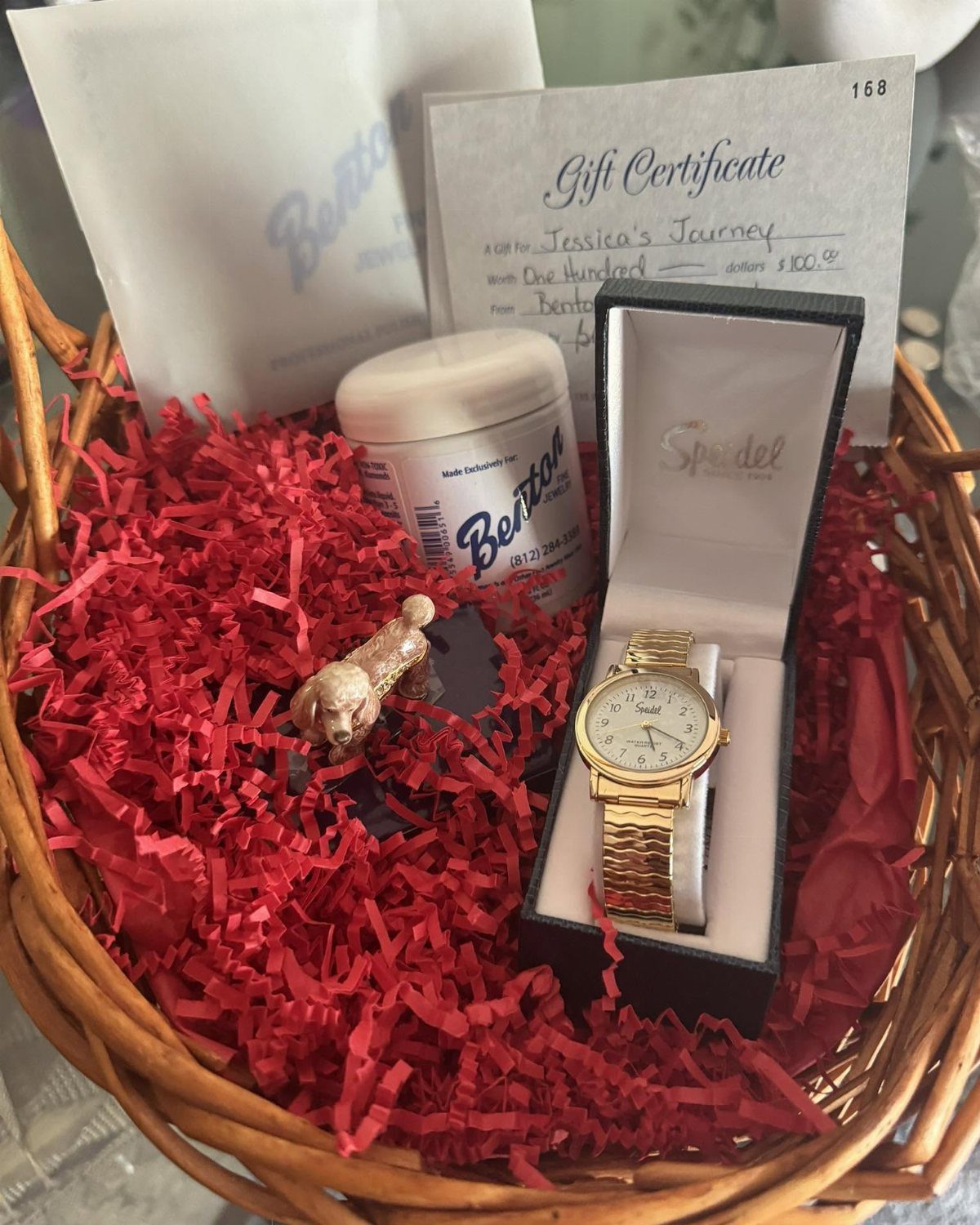 Wicker gift basket with red filler, gold watch, gift certificate, and cosmetic jar.