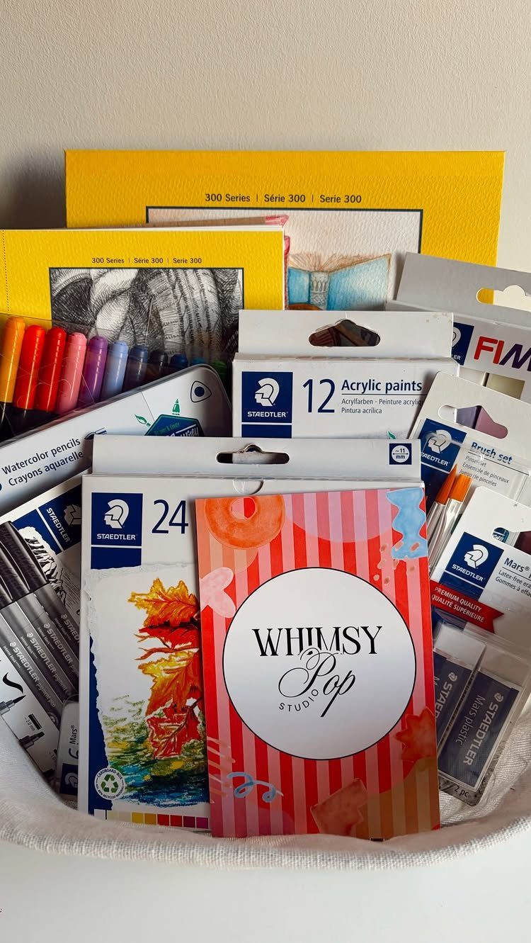 White gift basket filled with acrylic paints, watercolor pencils, markers, and sketch pads.