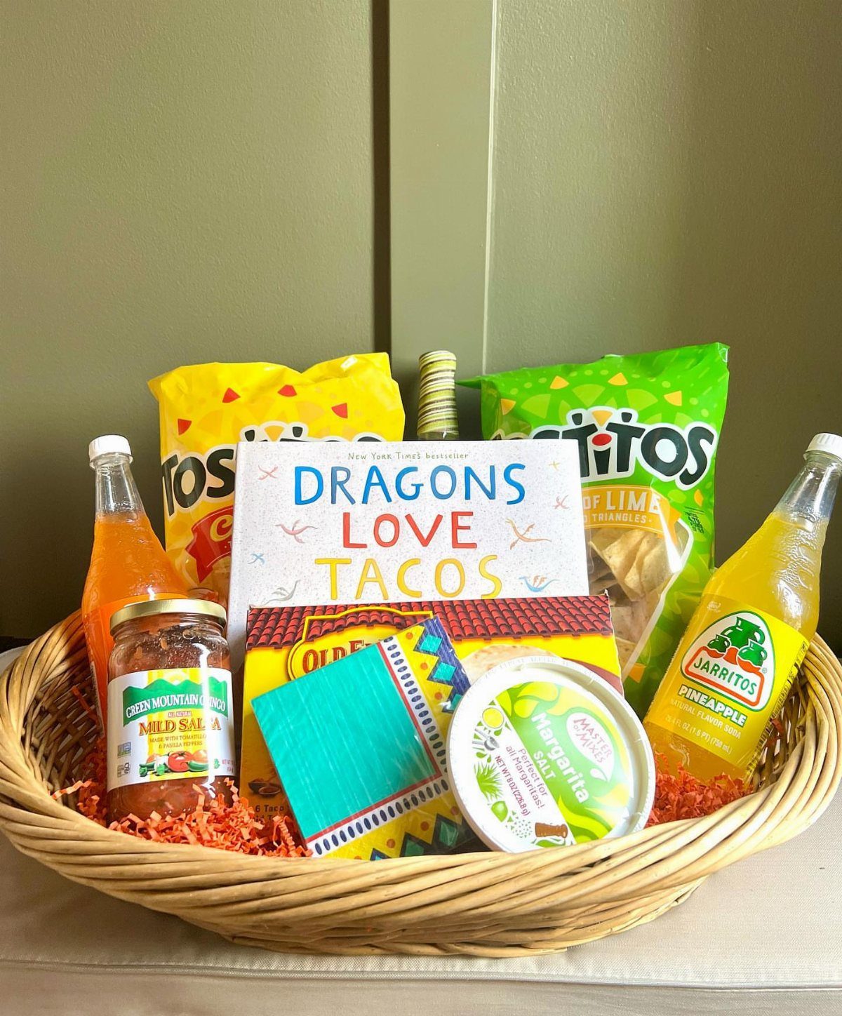 Wicker gift basket with taco-themed snacks, a children's book, and colorful sodas.