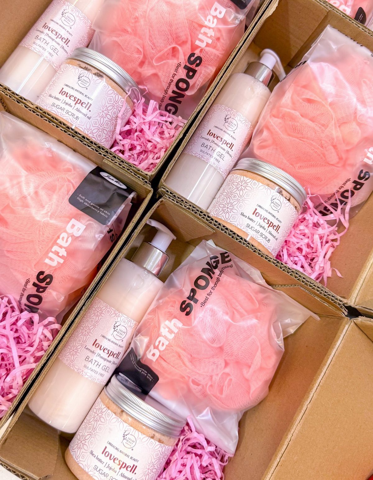 Three gift boxes containing pink bath sponges, bath gel, and sugar scrub jars.