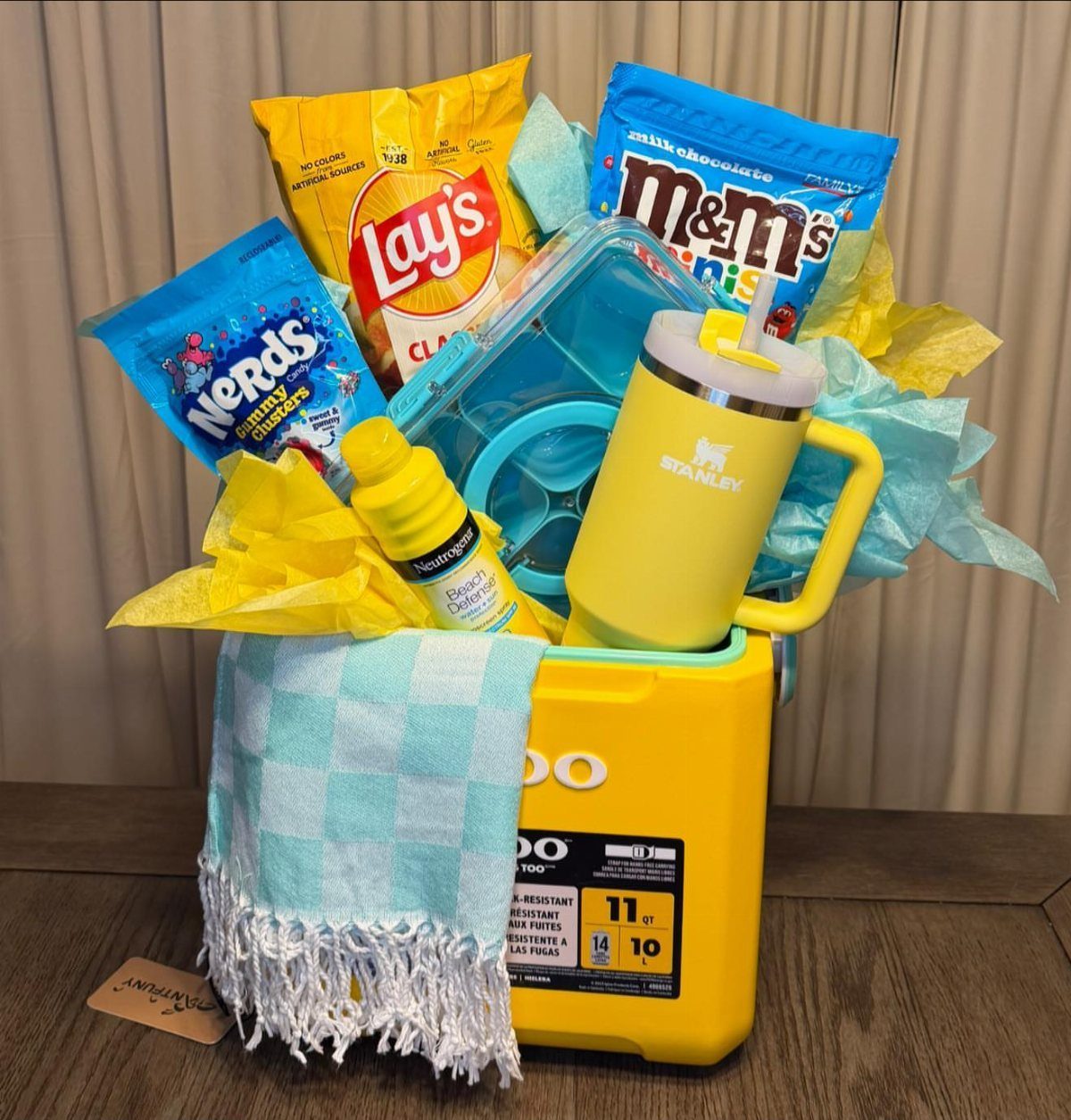 Yellow cooler raffle basket with snacks, a Stanley tumbler, and a blue towel.