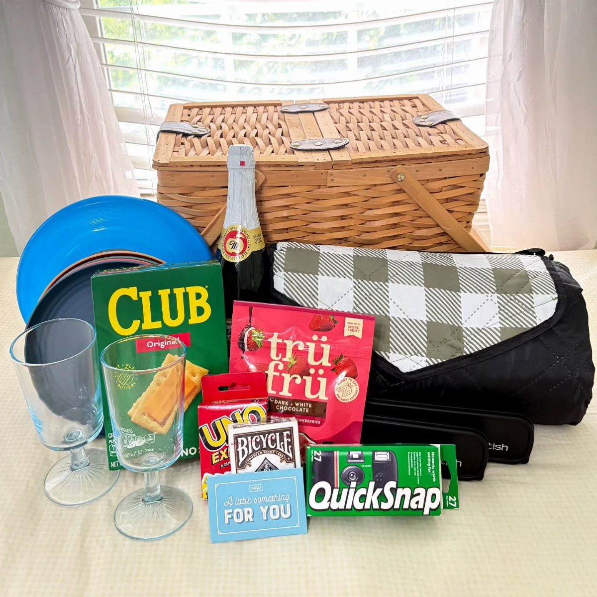 Wicker picnic hamper surrounded by snacks, drinks, and games on a white tablecloth.