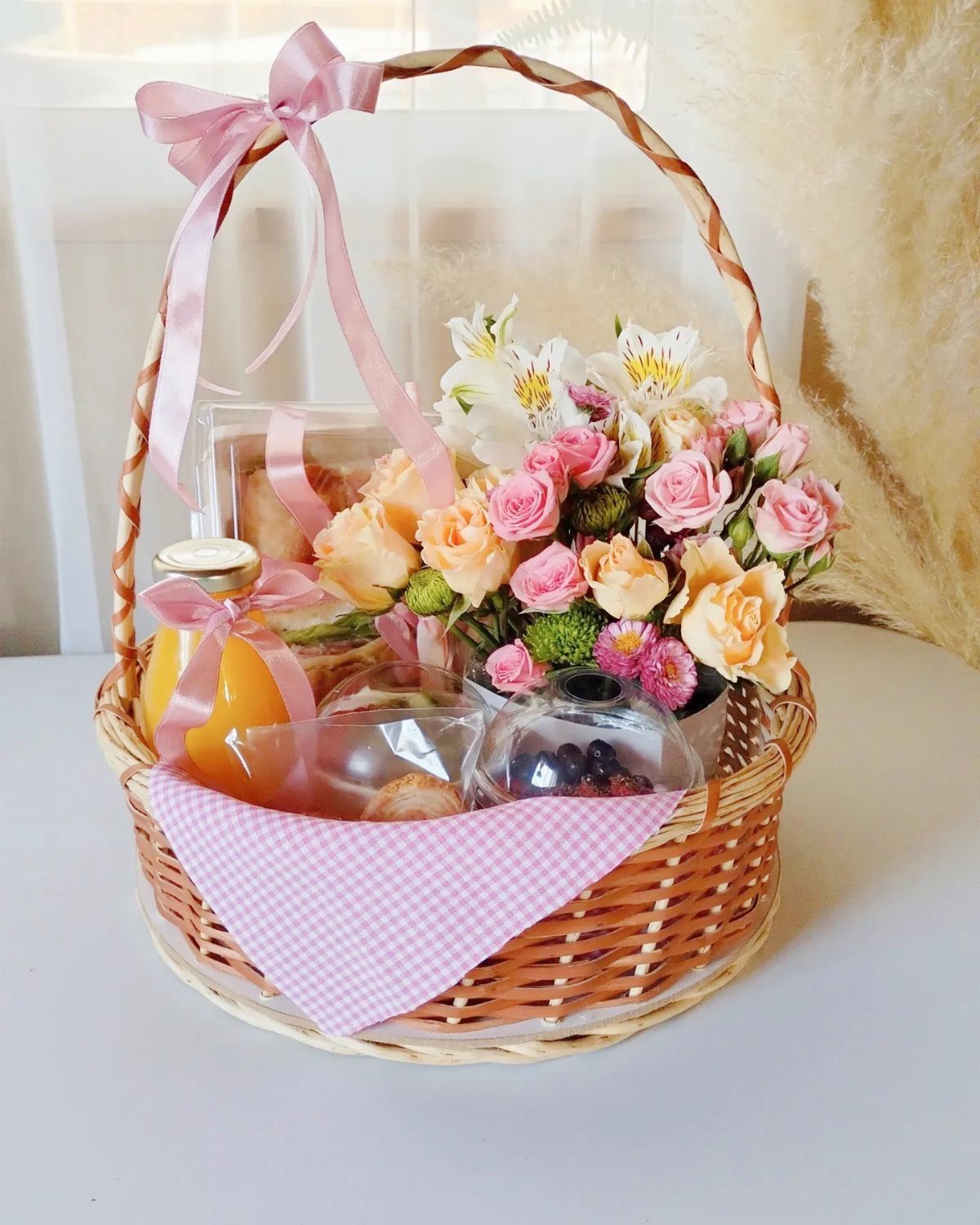 Wicker gift basket with orange juice, fresh roses, and pastries on a white surface.