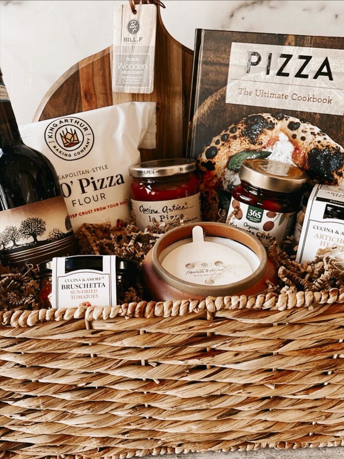 Woven gift basket containing pizza flour, sauce, a cookbook, and a wooden peel.