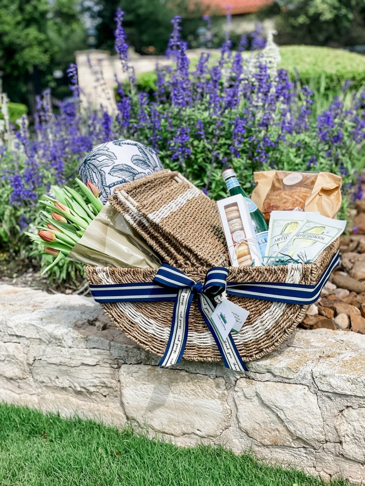 Woven picnic basket with blue ribbon, snacks, and tulips on a stone wall.