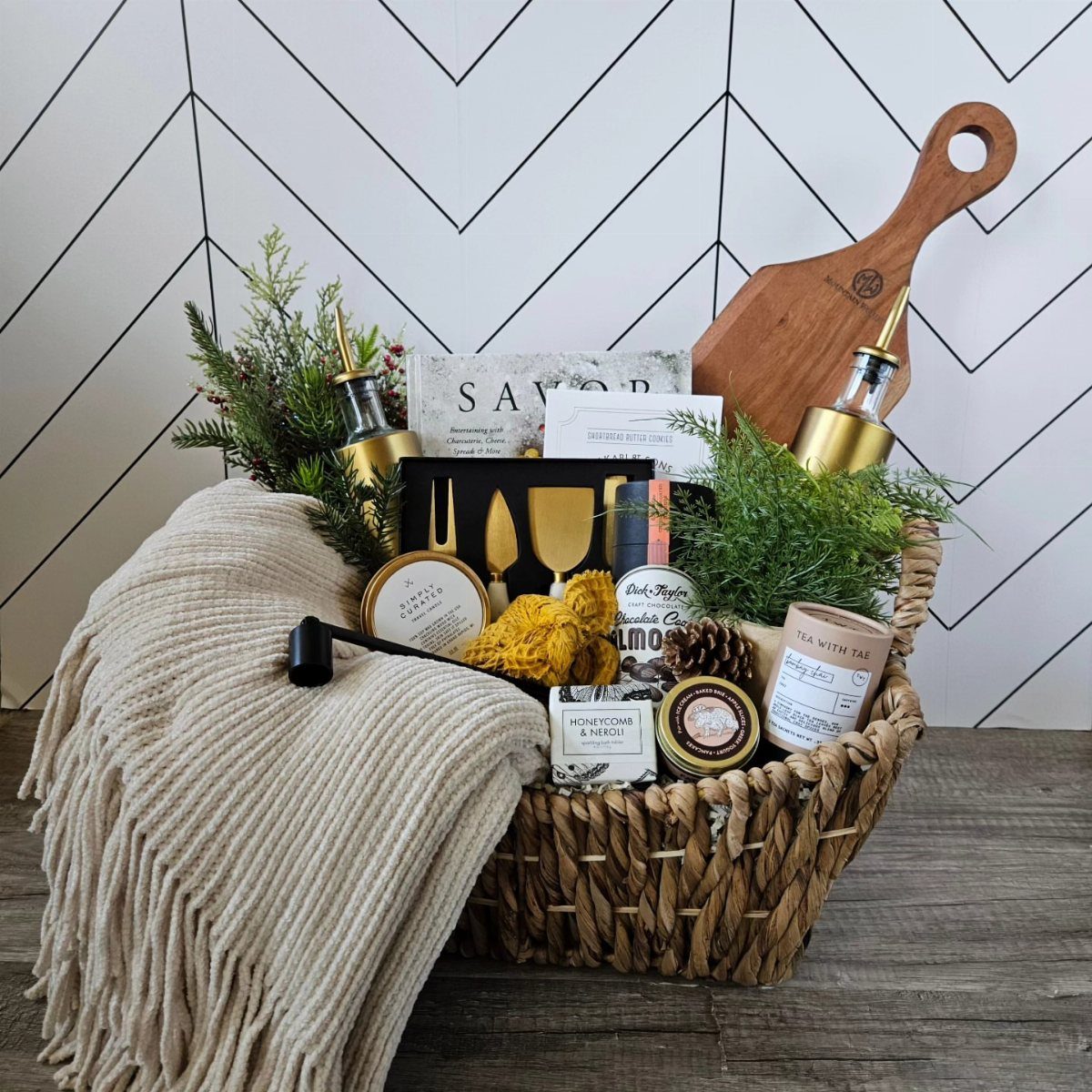 Woven gift basket with gourmet treats, a wooden board, and a beige throw blanket.