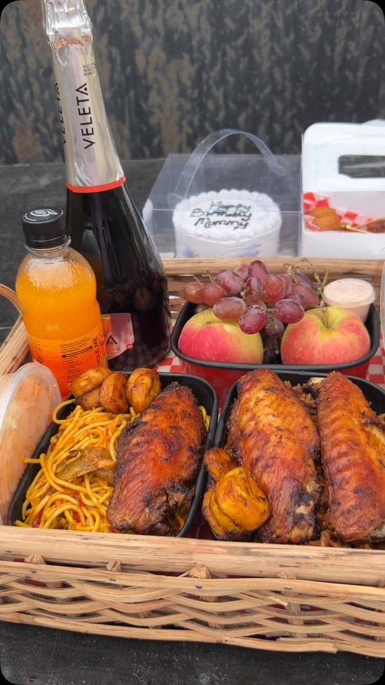 Wicker gift basket containing sparkling wine, orange juice, roasted chicken, pasta, and fruit.