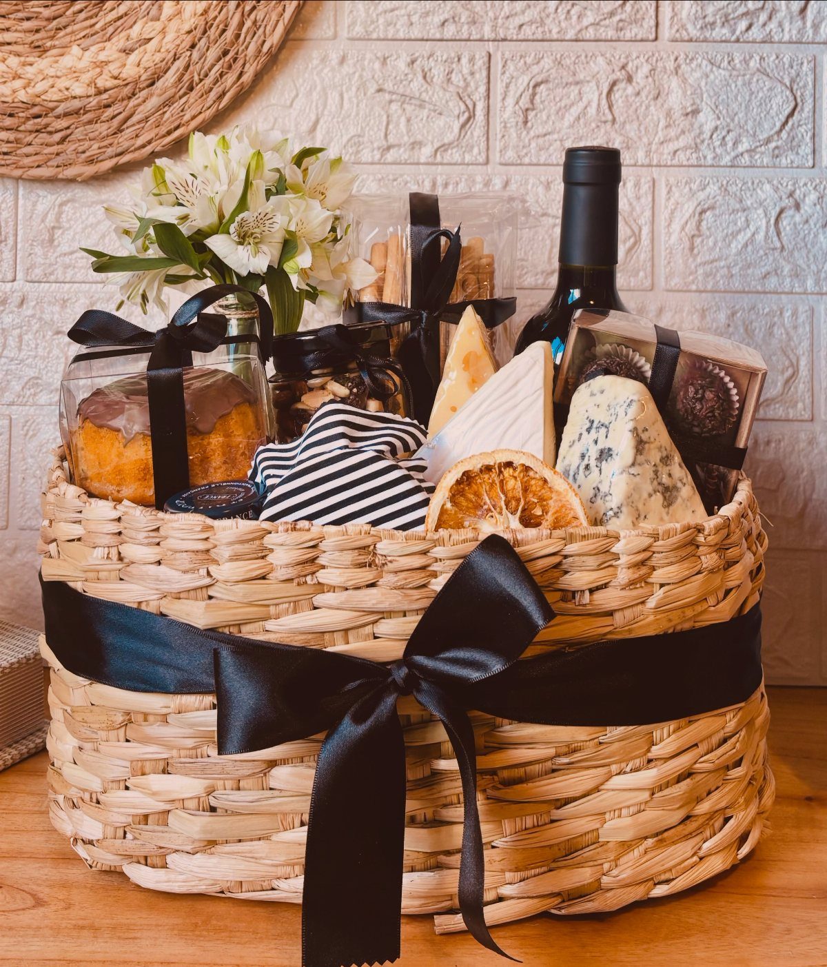 Woven gift basket with red wine, cheese wedges, cake, and white flowers.